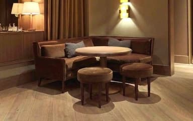 An L-shaped corner booth seating unit upholstered in a brown, velvet-like fabric, accompanied by a round dining table with a pedestal base and two matching upholstered stools. The seating features a high backrest and includes two small gray accent pillows. The table has a light wood finish, and all seating units have dark, tapered wooden legs. — 3D model
