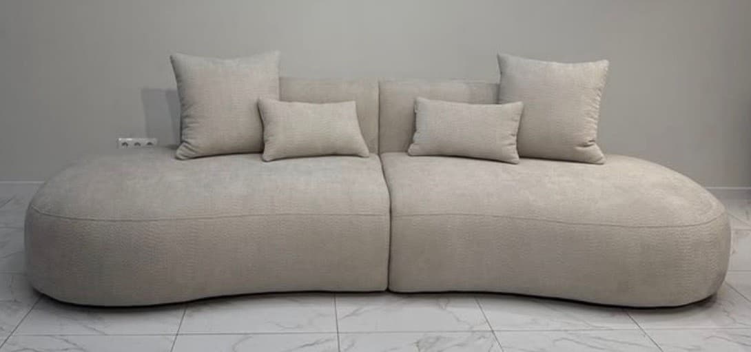 A light beige, modular sectional sofa with an organic, curvilinear form, composed of two distinct sections. It features plump, rounded edges and is upholstered in a textured fabric. The sofa includes multiple matching loose back cushions of varying sizes, contributing to its comfortable and inviting appearance. The design emphasizes softness and fluid lines, typical of modern and contemporary aesthetics. — 3D model