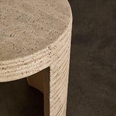 A cylindrical end table with a textured, stone-like surface. The table has a rounded top and a single, curved leg or base element visible. The side of the table exhibits a series of horizontal grooves or ridges, suggesting a layered construction or decorative carving. The material appears porous and natural, consistent with travertine or a similar stone composite. The color is a light, earthy beige. — 3D model