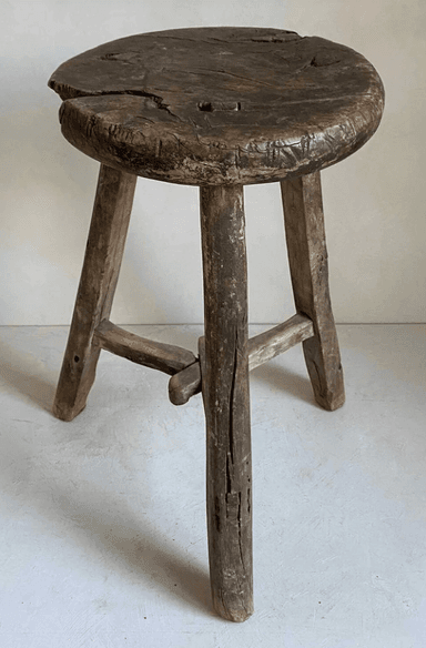 A rustic, round-top stool with three legs. The stool is constructed from weathered wood and shows signs of age, including cracks and a worn finish. The legs are roughly shaped and attached to the seat, with an additional wooden brace connecting two of the legs near the bottom. — 3D model
