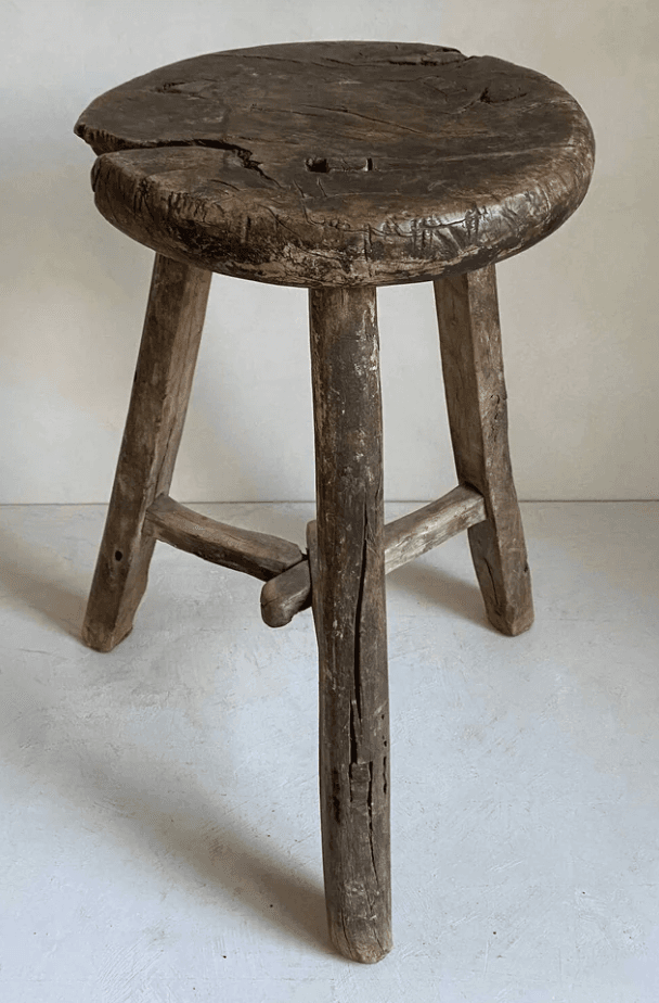 A rustic, round-top stool with three legs. The stool is constructed from weathered wood and shows signs of age, including cracks and a worn finish. The legs are roughly shaped and attached to the seat, with an additional wooden brace connecting two of the legs near the bottom. — 3D model