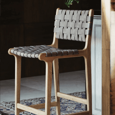 A bar stool with a light-colored wooden frame and woven leather straps for the seat and backrest. The wooden frame has a smooth, curved design, and the legs are connected by stretchers for stability. The leather straps are interlaced to form a textured surface for seating and back support. A patterned rug is visible beneath the stool. — 3D model