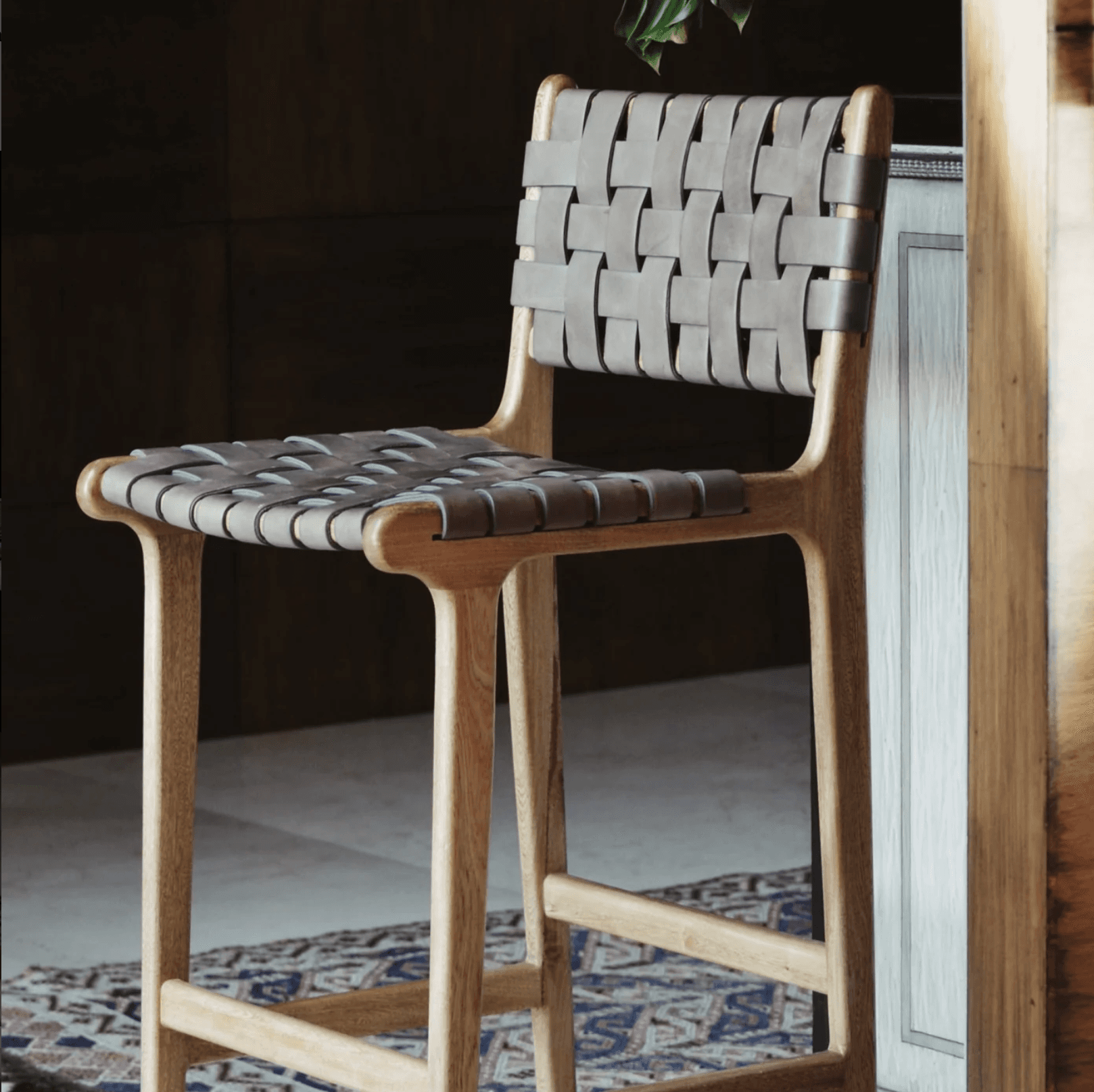 A bar stool with a light-colored wooden frame and woven leather straps for the seat and backrest. The wooden frame has a smooth, curved design, and the legs are connected by stretchers for stability. The leather straps are interlaced to form a textured surface for seating and back support. A patterned rug is visible beneath the stool. — 3D model