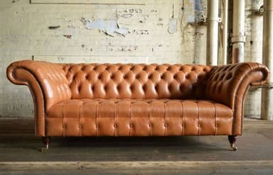 A classic Chesterfield sofa upholstered in tan leather, featuring deep button tufting across the backrest, seat, and rolled arms. The sofa has distinctive rolled armrests, decorative nailhead trim along the base and arms, and stands on short, turned legs with brass caster wheels. — 3D model
