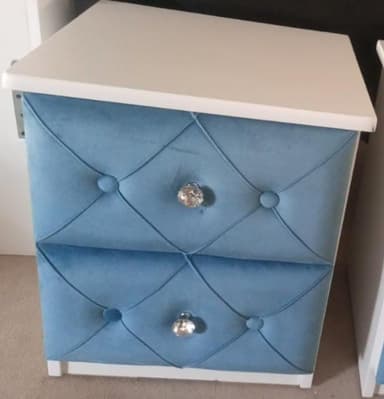 A compact two-drawer nightstand featuring a white frame and a white tabletop. The front of each drawer is upholstered in a diamond-tufted blue fabric, adorned with clear, faceted crystal-style knobs. The overall design suggests a contemporary or glam aesthetic. — 3D model