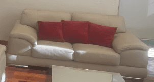 A modern two-seater sofa upholstered in light beige material, featuring three red throw pillows. It has gently rounded arms and a tailored back, supported by visible chrome-finished feet. — 3D model