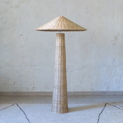 A tall, freestanding floor lamp constructed entirely from woven rattan or wicker. It features a slender, tapered cylindrical base that widens slightly towards the bottom for stability. The lamp is topped with a wide, shallow, conical shade, reminiscent of a mushroom cap, providing diffused downward light. The natural material gives it a bohemian or coastal aesthetic. — 3D model