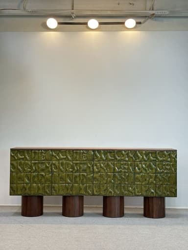 A long, low-profile credenza featuring a dark wood grain finish on its top and side panels, contrasting with a vibrant textured green front facade. The front is composed of square-like panels with an organic, wavy, or sculpted surface pattern, giving it a distinctive tactile appearance. The credenza is supported by four robust, cylindrical wood legs that match the main body's wood tone. — 3D model