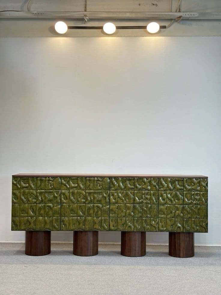 A long, low-profile credenza featuring a dark wood grain finish on its top and side panels, contrasting with a vibrant textured green front facade. The front is composed of square-like panels with an organic, wavy, or sculpted surface pattern, giving it a distinctive tactile appearance. The credenza is supported by four robust, cylindrical wood legs that match the main body's wood tone. — 3D model