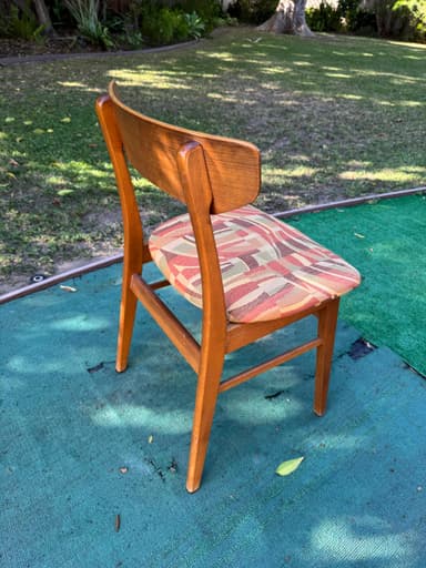 A close-up, overhead view of the padded seat and upper back support of an upholstered chair. The seat is covered in a multi-colored woven fabric featuring an abstract geometric pattern of wavy lines and rectangular blocks in shades of red, pink, beige, light green, and brown. Portions of the chair's wooden frame, including parts of the legs and back support, are visible, suggesting a medium-toned wood construction. The chair is positioned on a worn, teal-colored outdoor carpet or mat. — 3D model