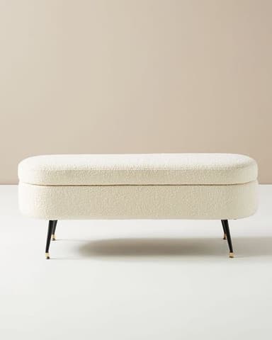 An oval-shaped, off-white boucle upholstered storage bench with slender black metal legs featuring brass tips. The top functions as a hinged lid, revealing internal storage. — 3D model