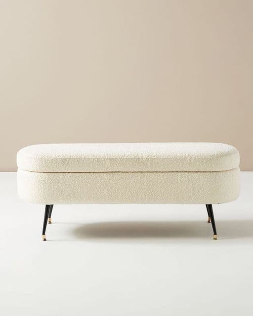An oval-shaped, off-white boucle upholstered storage bench with slender black metal legs featuring brass tips. The top functions as a hinged lid, revealing internal storage. — 3D model