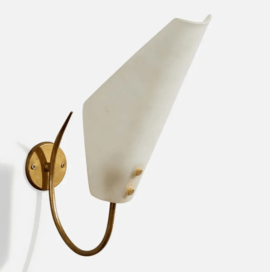 A vintage wall lamp featuring a conical white shade made of frosted or opaque glass, suspended from a curved brass arm. The base is a round brass plate designed for wall mounting, with a decorative, curved metal piece extending upwards from it. The shade is secured with two small brass finials. — 3D model
