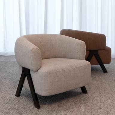 A modern armchair with a rounded, upholstered body and angled legs. The armchair features a curved backrest that flows into armrests, all covered in a textured light beige fabric. The base consists of two dark, V-shaped legs that are angled outwards, providing a stable support. The armchair is placed on a textured grey carpet. — 3D model