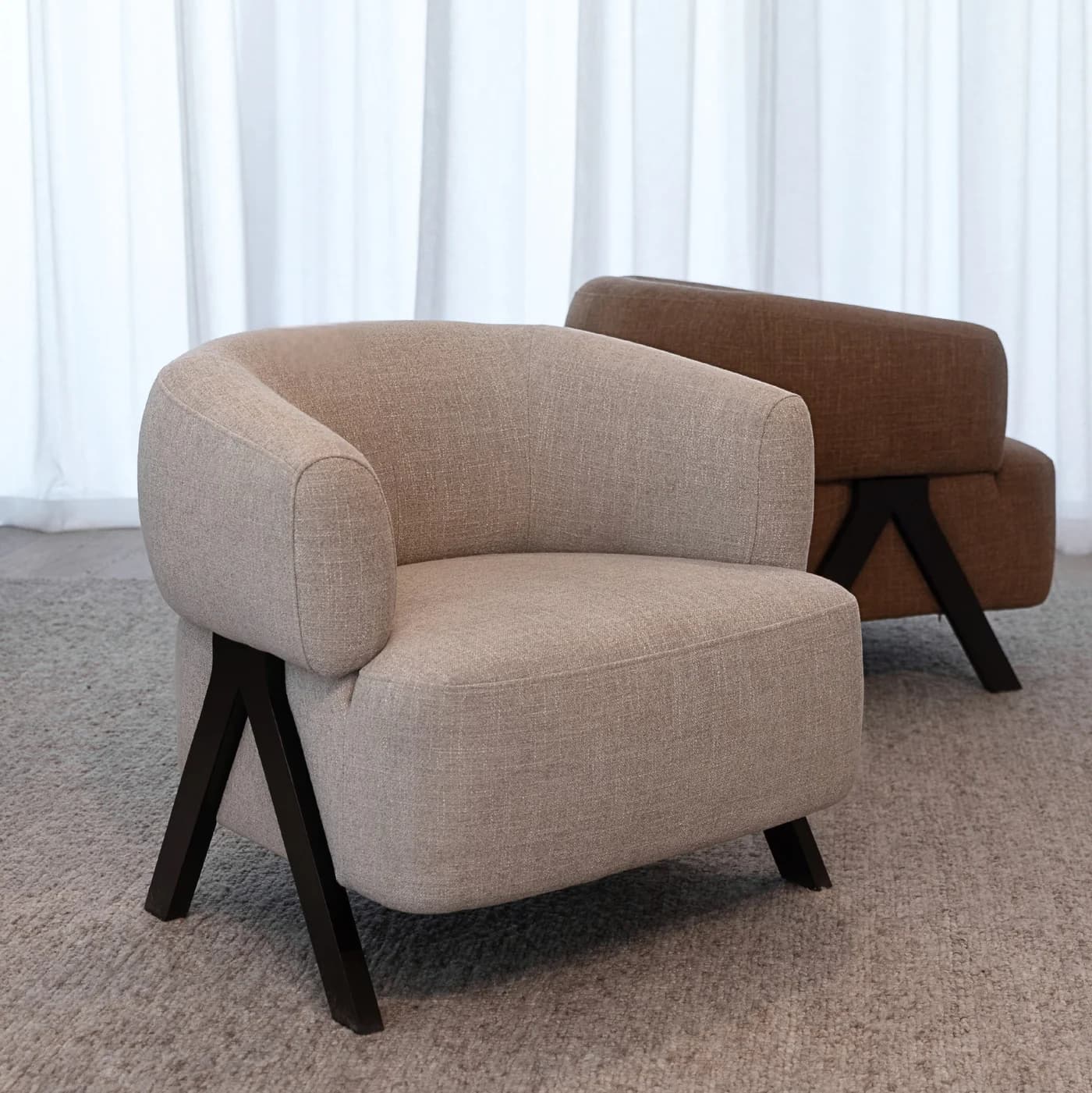 A modern armchair with a rounded, upholstered body and angled legs. The armchair features a curved backrest that flows into armrests, all covered in a textured light beige fabric. The base consists of two dark, V-shaped legs that are angled outwards, providing a stable support. The armchair is placed on a textured grey carpet. — 3D model