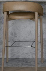 A modern bar stool featuring a round upholstered seat and a continuous, curved upholstered backrest that flows into integrated armrests. The frame consists of four slender, straight legs, likely metal, with a matching beige/taupe finish. A silver-toned metal footrest composed of interconnected bars provides support towards the bottom of the legs. The overall design is sleek and contemporary. — 3D model