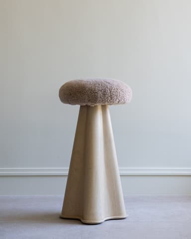 A tall, modern pedestal stool featuring a round, fluffy boucle upholstered seat in a light beige color, supported by a sculptural, flared wooden base with an organic, undulating silhouette. The base is crafted from light-toned wood with visible grain, tapering upwards to meet the seat and widening towards a subtly curved bottom edge. — 3D model