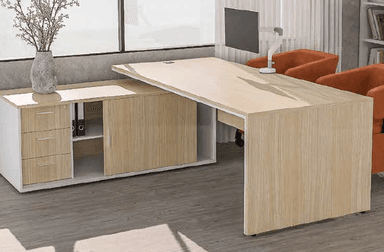 An L-shaped office desk featuring a spacious primary work surface in a light wood-tone laminate, supported by a panel leg. The L-extension is formed by an integrated storage unit, also in light wood laminate and white, which includes a three-drawer unit on one side and a sliding door cabinet on the other, with an open shelf section in between. A cable management grommet is visible on the main desk surface. — 3D model
