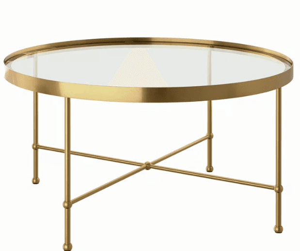 A modern round coffee table featuring a clear glass tabletop set within a polished brass-finished metal frame. The table stands on four slender, tapered legs connected by an X-shaped stretcher near the bottom, with small spherical decorative accents where the legs meet the stretcher and at the feet. — 3D model
