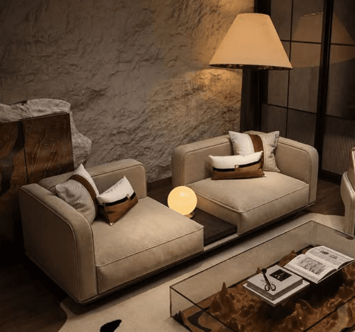 A modern modular sectional sofa featuring two upholstered seating units separated by a dark wood integrated side table. The sofa is upholstered in a light beige fabric, presenting a clean, contemporary aesthetic with rounded edges on the armrests and backrests. The integrated side table is made of dark wood, housing a spherical, illuminated lamp. Several decorative throw pillows with brown, white, and black accents are placed on the seating units. — 3D model