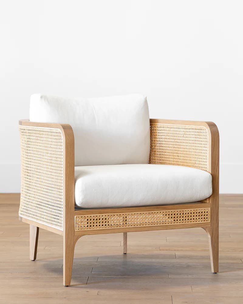 A wooden armchair with a cane woven back and sides, featuring a plush white seat and back cushion. The frame is light-colored wood with a natural finish. The legs are tapered and angled outwards. The cane pattern is a hexagonal weave. — 3D model