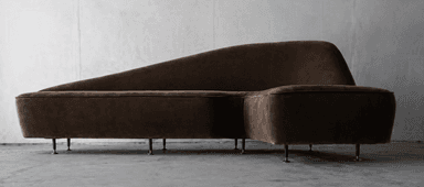 An L-shaped modular sectional sofa featuring a distinctively curved backrest on one side and an integrated chaise extension. The sofa is upholstered in a plush, dark brown fabric, likely velvet or microsuede, and supported by multiple small, dark, low-profile cylindrical feet. — 3D model