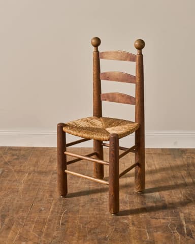 A rustic wooden side chair featuring a distinctive ladder back with three horizontal slats, a woven rush seat, and turned finials on the top of the back posts. The frame is constructed from warm-toned wood, likely oak or pine, with round, robust legs connected by simple stretchers. The overall aesthetic suggests a traditional or farmhouse style. — 3D model
