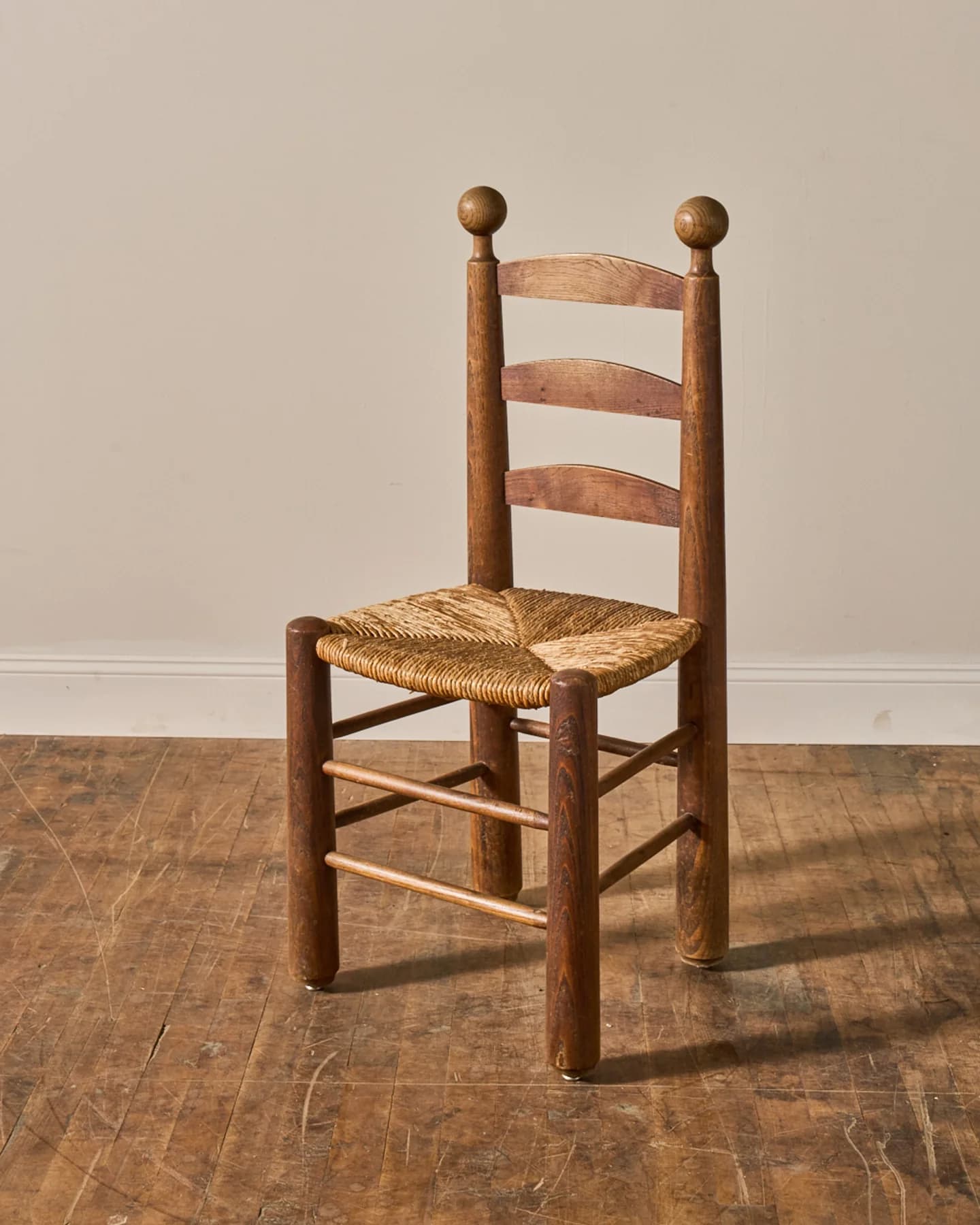 A rustic wooden side chair featuring a distinctive ladder back with three horizontal slats, a woven rush seat, and turned finials on the top of the back posts. The frame is constructed from warm-toned wood, likely oak or pine, with round, robust legs connected by simple stretchers. The overall aesthetic suggests a traditional or farmhouse style. — 3D model