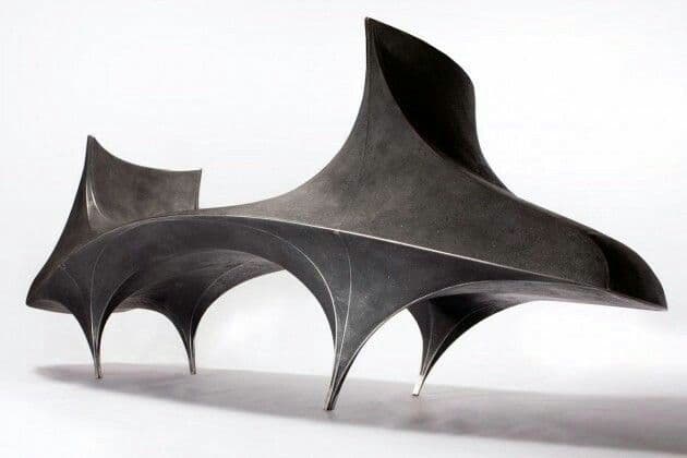 A sculptural bench with an organic, flowing form. The bench features a dark, matte finish and appears to be constructed from a molded material. It has a unique, futuristic aesthetic with dynamic curves and sharp angles. The base is formed by several pointed legs that create arched openings underneath. — 3D model