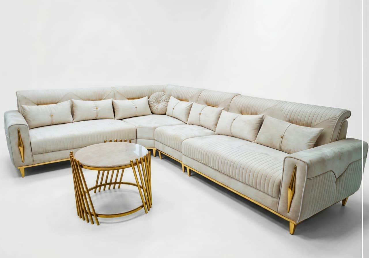An L-shaped contemporary sectional sofa upholstered in a light cream or beige fabric, featuring distinct gold-toned metal accents on its arms and legs. The sofa has a low profile, plush seat cushions, and multiple decorative throw pillows. The backrests and sides exhibit channel tufting and diamond-patterned quilting in some areas, adding textural interest and a luxurious aesthetic. The overall design suggests a blend of modern sophistication and glam elements. — 3D model