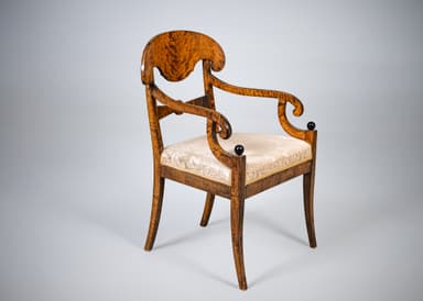 An antique armchair featuring a distinctive curved and carved backrest, likely made from curly maple or birch wood with a warm, amber finish. The chair has an upholstered seat covered in a cream-colored damask fabric with a patterned motif. The back legs are straight and square-tapered, and the back structure includes carved elements that function as armrest supports. The overall design suggests a formal, historical aesthetic. — 3D model