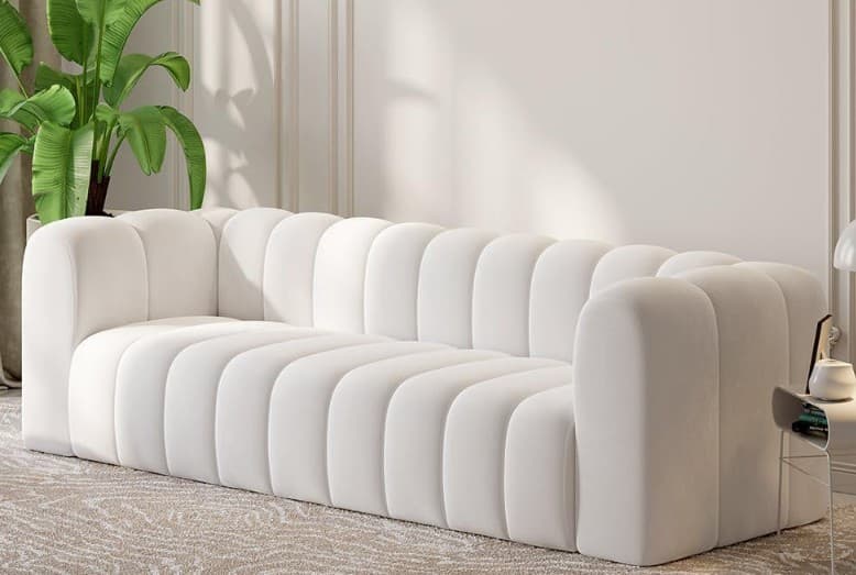 A long, contemporary sofa featuring distinctive channel tufting across its backrest and seat, creating a plush, ribbed appearance. The sofa has thick, rounded armrests that flow seamlessly from the back, and it sits low to the ground with no visible legs, emphasizing a solid, monolithic form. Upholstered in a soft, white or off-white fabric, likely velvet or boucle, it presents a luxurious and inviting aesthetic. — 3D model