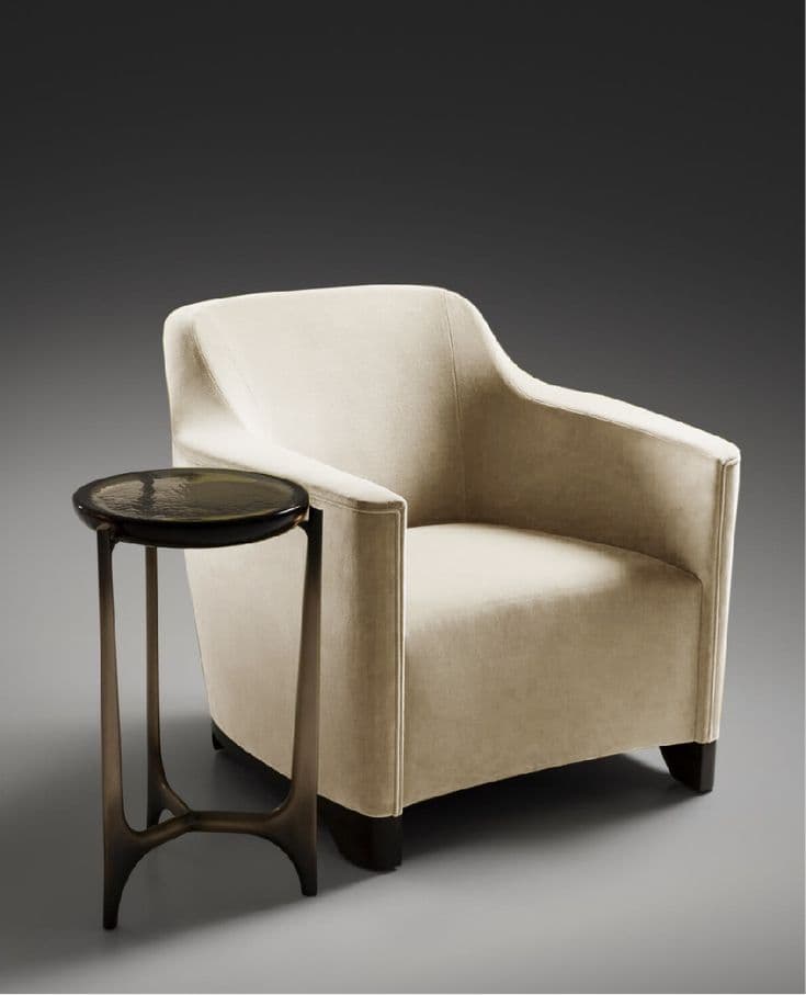 A single upholstered accent chair with a rounded back and armrests, accompanied by a small, dark, round-top side table with a sculptural base. The chair is upholstered in a textured fabric and has dark legs. The side table features a circular top with a textured surface and a dark, metallic-looking base with three legs that converge towards the center. — 3D model