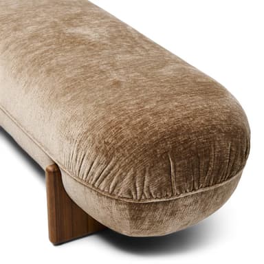 A low-profile, elongated bench with a rounded, upholstered form and solid wood legs. The upholstery features a textured, velvet-like fabric in a neutral tone, exhibiting subtle creasing that follows the rounded shape. The legs are triangular in cross-section and appear to be made of a dark wood with a visible grain. — 3D model