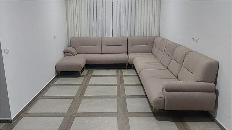 A large L-shaped modular sectional sofa in a light beige fabric, featuring plush upholstered cushions and backrests with horizontal stitch detailing. The sofa is composed of multiple individual units, including a corner piece, armless sections, and an end piece with an armrest. It is supported by short, tapered wooden legs. An accompanying rectangular ottoman matches the sofa's upholstery and leg design. — 3D model