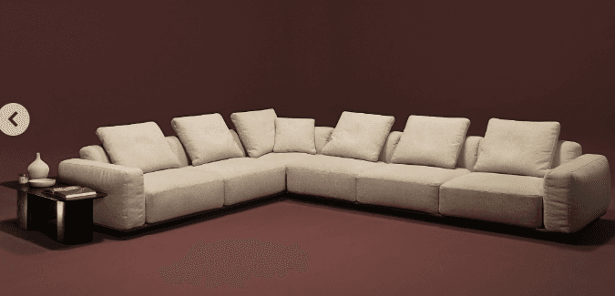 An L-shaped modular sectional sofa, upholstered in a light beige fabric, featuring a minimalist design with a low-profile, recessed dark plinth base. The sofa is composed of multiple individual units arranged in a corner configuration, providing extensive seating. It includes numerous loose back cushions and throw pillows, enhancing comfort and aesthetic appeal. The overall appearance is modern and clean-lined. — 3D model