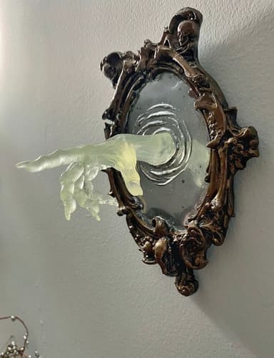This decorative wall mirror features an elaborately carved, antique-style frame with gothic-inspired motifs, including skulls. Emerging from the mirrored surface is a translucent, sculpted hand, rendered in a pale, ghostly hue, creating a surreal and artistic statement piece. The mirror itself has a light, perhaps frosted or etched, surface that offers a soft reflection. — 3D model