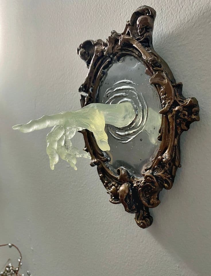 This decorative wall mirror features an elaborately carved, antique-style frame with gothic-inspired motifs, including skulls. Emerging from the mirrored surface is a translucent, sculpted hand, rendered in a pale, ghostly hue, creating a surreal and artistic statement piece. The mirror itself has a light, perhaps frosted or etched, surface that offers a soft reflection. — 3D model