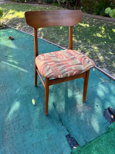 A close-up, overhead view of the padded seat and upper back support of an upholstered chair. The seat is covered in a multi-colored woven fabric featuring an abstract geometric pattern of wavy lines and rectangular blocks in shades of red, pink, beige, light green, and brown. Portions of the chair's wooden frame, including parts of the legs and back support, are visible, suggesting a medium-toned wood construction. The chair is positioned on a worn, teal-colored outdoor carpet or mat. — 3D model