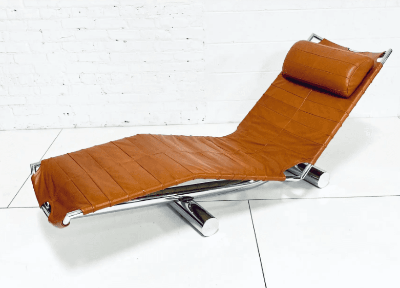 A contemporary chaise lounge with a curved, reclining design. The seat and back are upholstered in a stitched, padded orange-brown leather or leather-like material. The frame appears to be constructed from polished chrome or stainless steel, providing a sleek, modern aesthetic. A cylindrical headrest pillow, matching the upholstery, is attached to the upper section. — 3D model