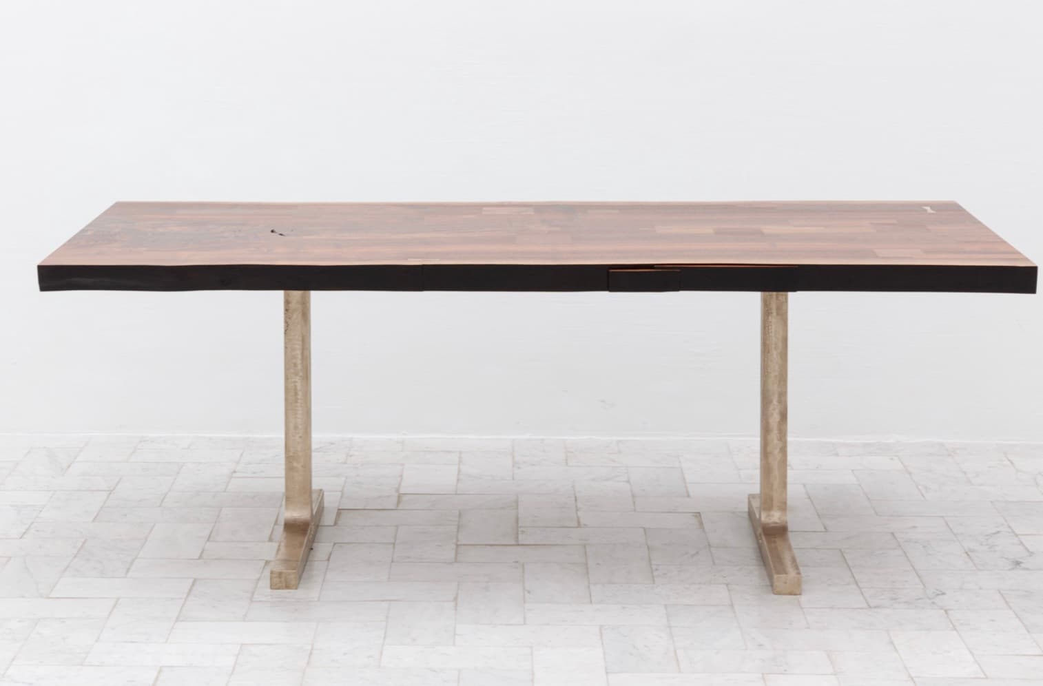 A rectangular dining table with a thick, butcher block style wooden top. The top is constructed from numerous rectangular wood pieces arranged in a parquet pattern. The edges of the tabletop appear to have a live-edge or rough-hewn finish on the longer sides. It is supported by two metal legs, each with a T-shaped base. The legs and bases have a metallic, possibly brushed or aged finish. A small rectangular drawer or insert is visible on the front edge of the table. — 3D model
