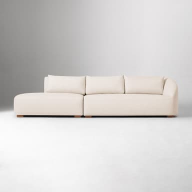 A sectional sofa with a chaise lounge. The sofa has a low profile and is upholstered in a textured fabric. It features plush back cushions and a rounded armrest on the chaise end. The sofa is supported by small, dark wooden block feet. — 3D model