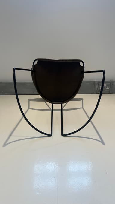 A contemporary armchair with a dark brown leather seat and backrest supported by a slender black metal frame. The frame features a curved base that extends into the legs, providing stability and a dynamic visual element. The seat and backrest are subtly contoured for comfort and appear to be constructed from stitched leather panels. — 3D model