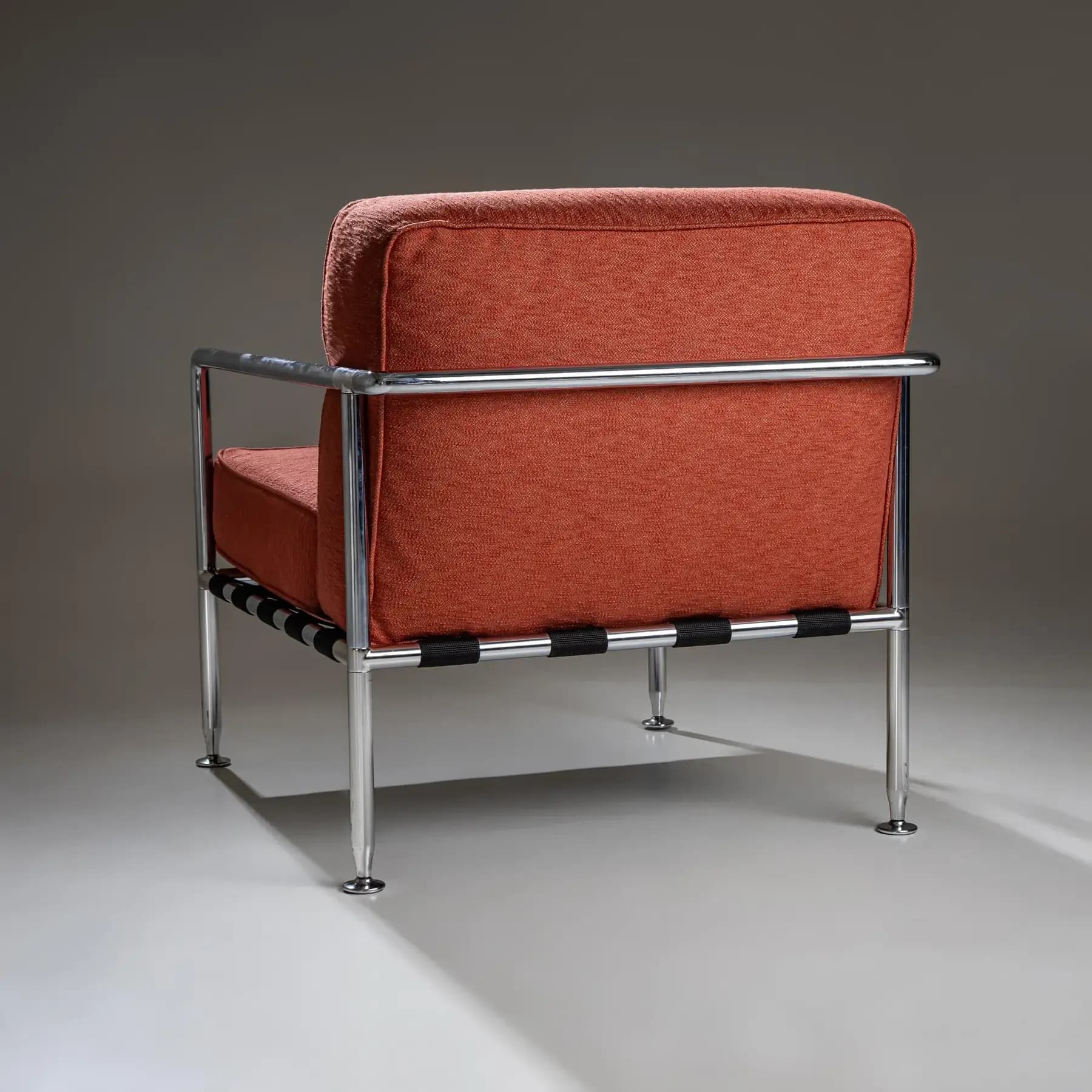 An armchair with a chrome-plated tubular steel frame and upholstered cushions. The seat and back cushions are covered in a textured, rust-colored fabric. The frame consists of slender legs, armrests, and a horizontal back support. Black woven straps are visible underneath the seat, connecting the frame elements. — 3D model