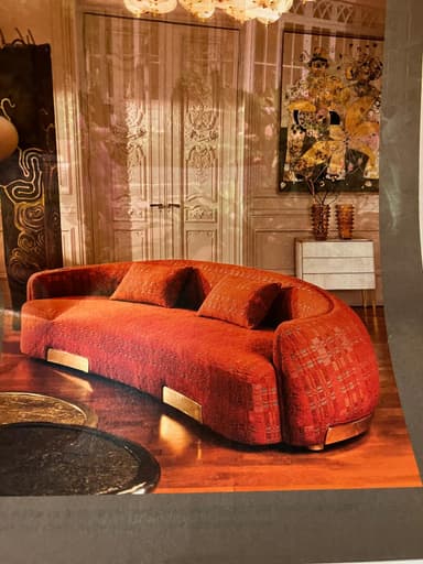 A contemporary, curved sofa with a textured, rust-orange upholstery. It features a subtle woven pattern within the fabric and is accented with polished brass trim at the base. Two matching throw pillows are included. — 3D model