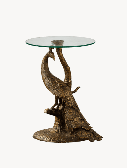 A decorative accent table featuring a base sculpted in the form of a peacock standing on a branch, supporting a circular glass tabletop. The peacock is intricately detailed with feathers and perched on a textured base. — 3D model