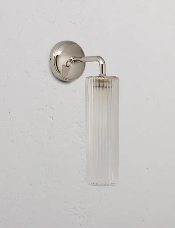 A modern wall sconce featuring a cylindrical ribbed glass shade attached to a polished metal arm and a circular wall mount. The glass shade has vertical fluting that diffuses light. The metal components appear to be polished nickel or a similar bright, reflective finish. The sconce is mounted on a textured white wall. — 3D model