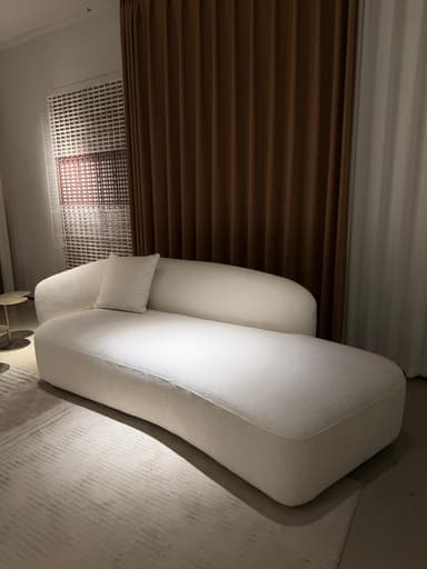 An asymmetrical upholstered chaise lounge in a cream-colored textured fabric, featuring a single integrated armrest/backrest and a curved, elongated form for reclining. — 3D model