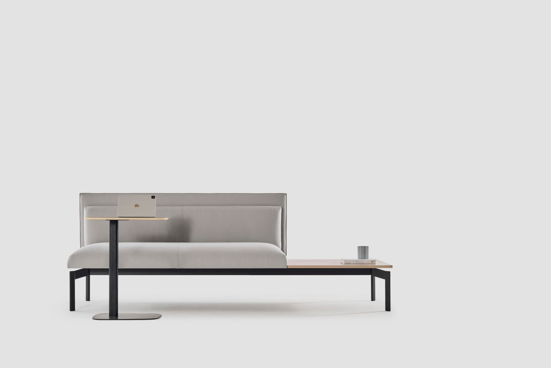 A modern upholstered bench with integrated side tables. The bench features a light gray fabric upholstery for the seat and backrest, with a subtle horizontal stitch detail on the back. On the left side, a black metal framed table with a light wood surface is attached, supporting a laptop. On the right side, a continuous light wood surface extends, serving as an integrated side table, on which a book and a cylindrical object are placed. The bench has a minimalist black metal frame and legs. — 3D model