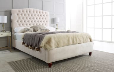 A cream-colored upholstered bed with a diamond-tufted headboard featuring crystal-like embellishments. The bed frame is also upholstered in a matching cream fabric. It is shown with a fluffy cream-colored throw blanket, two plaid throw pillows, and a gray knitted throw. The bed rests on four dark wooden carved legs. Two matching nightstands with drawers and brass accents are positioned on either side of the bed, each holding a white lampshade with a brass base. — 3D model